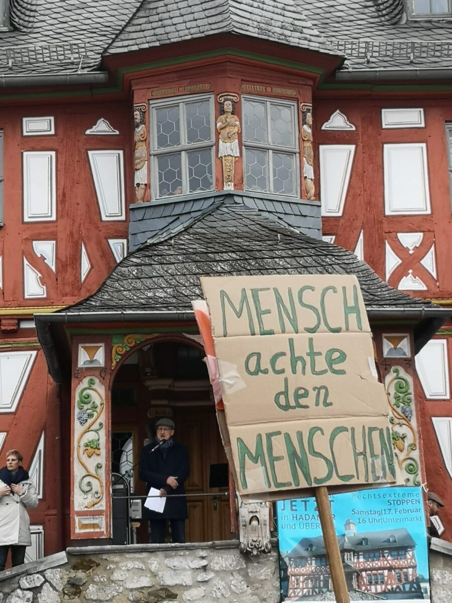 Ein Foto von einem Plakat auf einer Demonstration. Auf Pappkarton steht in grüner Schrift: "Mensch achte den Menschen". Im Hintergrund ist ein Fachwerkhaus zu sehen.